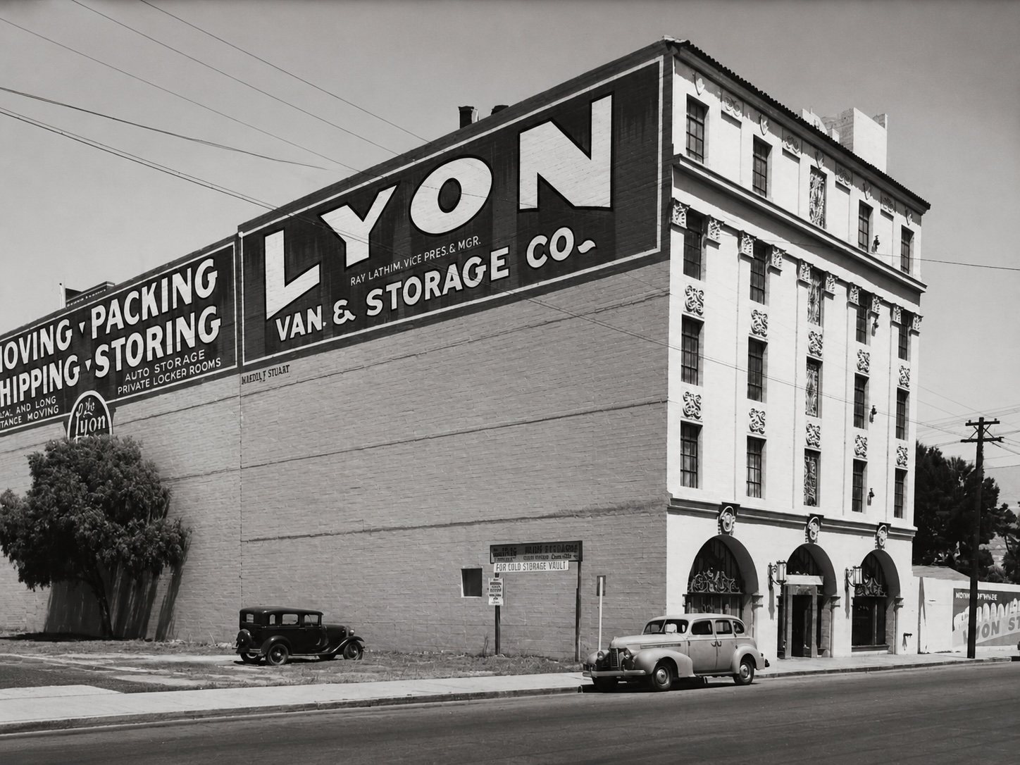 Historic photograph of the Lyon Van and Storage Co. building from Cota Street, its upper flank painted with a large sign reading Moving, Packing, Shipping, Storing, with period automobiles at the kerb.