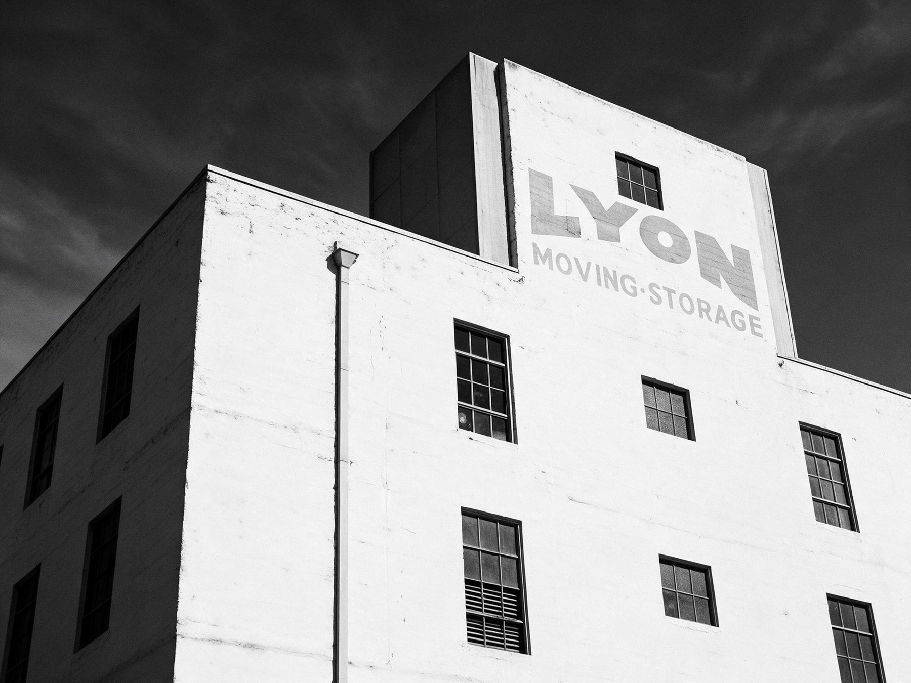 The upper storeys of the Lyon Building against a dark sky, the white flank wall carrying painted lettering reading LYON MOVING and STORAGE, with the blank frame of the former roof sign standing behind it.